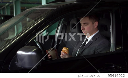A young businessman is having lunch in a car in a parking lot. A man in a suit eats fast food and enjoys the taste. The businessman eats a cheeseburger and drinks coffee. A young businessman is having lunch in a car in a parking lot. A man in a suit eats fast food and enjoys the taste. The businessman eats a cheeseburger and drinks coffee. 95416922