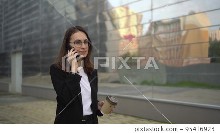 A young businesswoman is talking on the phone and drinking coffee walking around the business center. A woman in glasses and a suit is on her lunch break. A young businesswoman is talking on the phone and drinking coffee walking around the business center. A woman in glasses and a suit is on her lunch break. 95416923