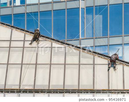 Two industrial climbers with safety tethers wash windows or repair walls of modern skyscraper. Dangerous work in the metropolis. Two industrial climbers with safety tethers wash windows or repair walls of modern skyscraper. Dangerous work in the metropolis. 95416971