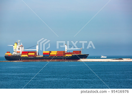 a bulk carrier with cranes for unloading cargo transports multi-colored containers, a small-sized sea vessel sails along the sea along the breakwater next to the port against the sky on a sunny day. a bulk carrier with cranes for unloading cargo transports multi-colored containers, a small-sized sea vessel sails along the sea along the breakwater next to the port against the sky on a sunny day. 95416976