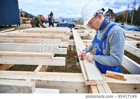 Man worker building wooden frame house on pile foundation. Carpenter using tape measure for measuring wooden planks and making marks with pencil. Carpentry concept. 95417149