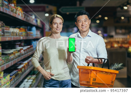 Young family diverse couple of shoppers in supermarket, smiling and looking at camera, grocery department, man and woman holding shopping basket and showing green screen of smartphone. 95417397