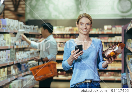 Portrait of a woman buyer in a supermarket in the grocery department, a blonde housewife chooses goods and makes purchases looking at the camera and smiling. Portrait of a woman buyer in a supermarket in the grocery department, a blonde housewife chooses goods and makes purchases looking at the camera and smiling. 95417403