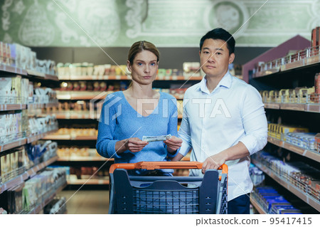 Upset young couple in supermarket, man and woman looking at camera poor have no opportunity and money to shop, multiracial family. 95417415
