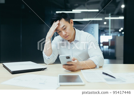 Confused Asian guy holding smart phone feels concerned thinking over received message. Mobile phone everyday usage, unpleasant news, waiting for important call, low signal device problems concept 95417446
