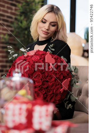 Smiling woman holding red roses bouquet, blurred giftboxes on table, valentines day luxury presents, romantic date. Excited blonde girlfriend sitting with flowers from boyfriend 95422746