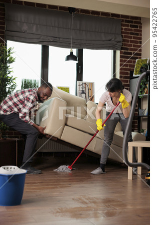 African american man lifts sofa while woman dusts underneath with mop, roommates doing spring cleaning in city apartment. Multiracial friends taking care of keeping home clean and tidy. 95422765