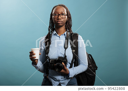 Serious looking professional photographer having DSLR device and cup of coffee while looking at camera on blue background. Young photography entusiast having photo equipment while enjoying beverage. 95422808