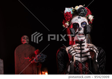 Creepy santa muerte model posing with black roses, acting like holy goddess of death on day of the dead mexican holiday. Celebrating halloween ritual with la cavalera catrina skull body art. 95423009