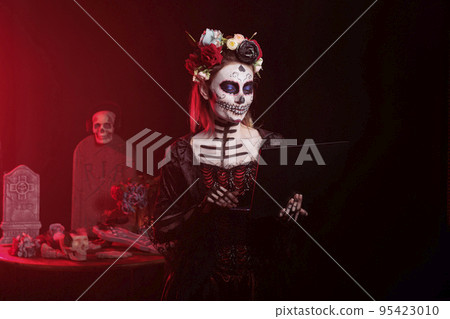 Halloween model using laptop in studio, looking like lady of the holy death or santa muerte. Wearing skull make up and cavalera catrina carnival costume, looking spooky and creepy with flowers. Halloween model using laptop in studio, looking like lady of the holy death or santa muerte. Wearing skull make up and cavalera catrina carnival costume, looking spooky and creepy with flowers. 95423010