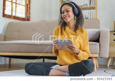Portrait of an African American with headphones and tablet smiling happily listen music while relaxing on the sofa at home. 95425505