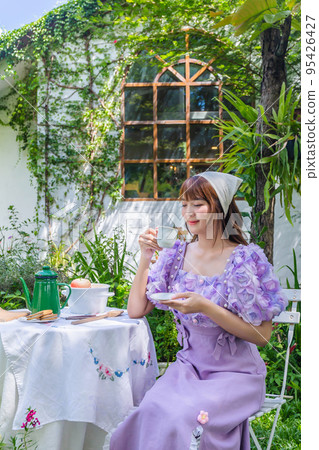 young woman in a princess dress sits drinking tea on a vintage table with cookies and books in a backyard with plants and flowers. young woman in a princess dress sits drinking tea on a vintage table with cookies and books in a backyard with plants and flowers. 95426427