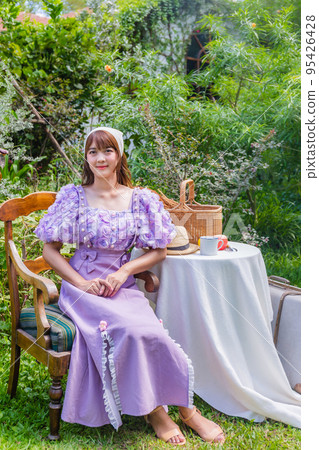 young woman in a princess dress sits drinking tea on a vintage table with cookies and books in a backyard with plants and flowers. young woman in a princess dress sits drinking tea on a vintage table with cookies and books in a backyard with plants and flowers. 95426428