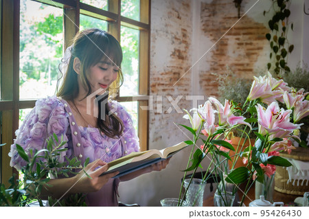 Girl in a princess dress reads a book in a room with windows and lots of green plants and lilies in a vase on the table like a fairy tale. Soft focus 95426430