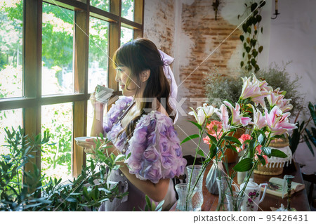 girl in a princess dress stands drinking tea at a window with plants, flowers and candles like in a fairy tale. Soft focus 95426431