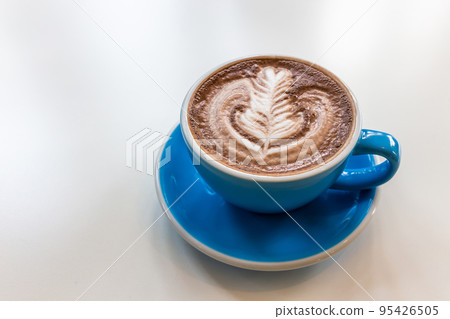 Hot cocoa in a blue cup on a white table with leaf motifs made of frothed milk. Hot cocoa in a blue cup on a white table with leaf motifs made of frothed milk. 95426505