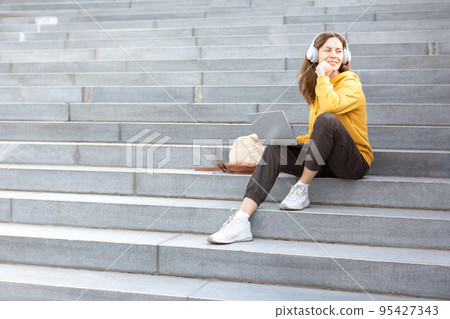 Student life, additional education. Adult caucasian woman in casual wear and headset sits on the steps of the stairs with a laptop outdoor and ponder something, selected focus. 95427343