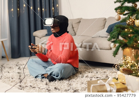 Muslim girl celebrating Christmas alone. Girl sitting near Christmas tree, wearing virtual reality glasses. Woman wearing rose sweater and black hijab. Muslim girl celebrating Christmas alone. Girl sitting near Christmas tree, wearing virtual reality glasses. Woman wearing rose sweater and black hijab. 95427779
