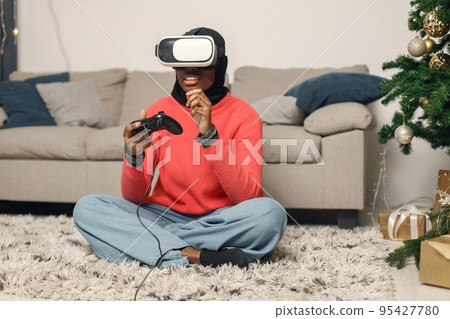 Muslim girl celebrating Christmas alone. Girl sitting near Christmas tree, wearing virtual reality glasses. Woman wearing rose sweater and black hijab. 95427780