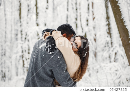 Indian man and caucasian woman walking in winter forest together. Lovely couple hugging. Man wearing grey coat and woman beige coat. 95427835