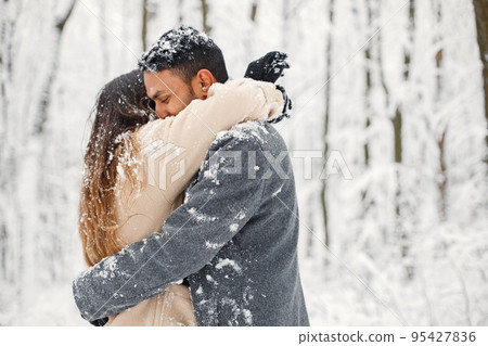 Indian man and caucasian woman walking in winter forest together. Lovely couple hugging. Man wearing grey coat and woman beige coat. 95427836