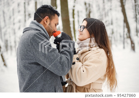 Indian man and caucasian woman walking in winter forest together. Lovely couple hugging and holding a thermos with hot tea. Man wearing grey coat and woman beige coat. Indian man and caucasian woman walking in winter forest together. Lovely couple hugging and holding a thermos with hot tea. Man wearing grey coat and woman beige coat. 95427860
