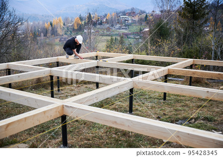 Man worker building wooden frame house on pile foundation. Carpenter measuring wooden planks and making marks with pencil. Carpentry concept. 95428345