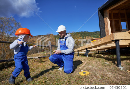 Father with toddler son building wooden frame house. Boy helping his daddy, playing with tape measure on construction site, wearing helmet and blue overalls on sunny day. Carpentry and family concept. 95430301