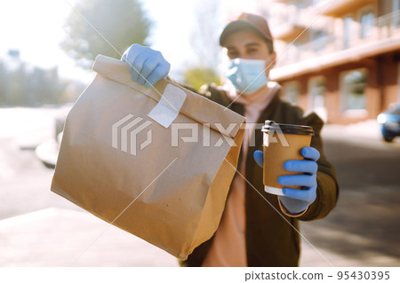 Courier in protective mask and medical gloves delivers takeaway food and coffee. Covid-19. Stay at home. Courier in protective mask and medical gloves delivers takeaway food and coffee. Covid-19. Stay at home. 95430395