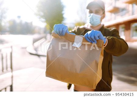 Courier in protective mask and medical gloves delivers takeaway food and coffee. Covid-19. Stay at home. 95430415
