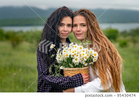 Two women enjoy nature in a field of daisies. Girlfriends hugging hold a bouquet of daisies and look at the camera. 95430474