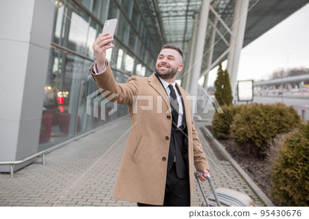young handsome businessman goes to the airport with a suitcase talks on the phone, smiles, calls a taxi, looking for. waiting for a meeting young handsome businessman goes to the airport with a suitcase talks on the phone, smiles, calls a taxi, looking for. waiting for a meeting 95430676