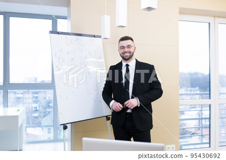 Young handsome businessman communicates, conducts video calls, conferences, meetings, works online, uses a laptop, writes on a flipchart, explains. Looks at the camera, smiles 95430692