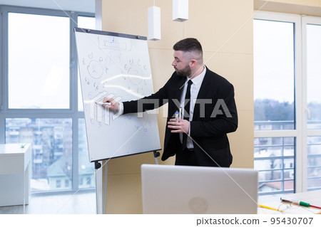 Young handsome businessman communicates, conducts video calls, conferences, meetings, works online, uses a laptop, writes on a flipchart, explains. Looks at the camera, smiles Young handsome businessman communicates, conducts video calls, conferences, meetings, works online, uses a laptop, writes on a flipchart, explains. Looks at the camera, smiles 95430707