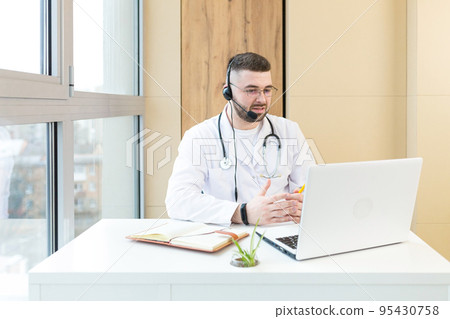 Young doctor man in headphones and with a microphone, advises on video communication of the patient, sits at the table, uses a laptop Young doctor man in headphones and with a microphone, advises on video communication of the patient, sits at the table, uses a laptop 95430758