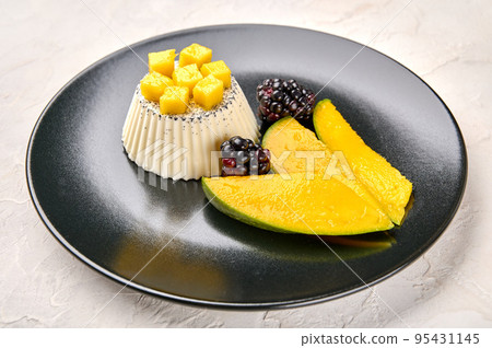 Close up panna cotta homemade with natural vanilla pod, with mango slices and blackberries on black Close up panna cotta homemade with natural vanilla pod, with mango slices and blackberries on black 95431145