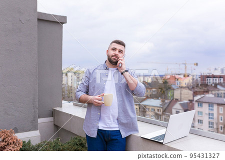 Young man working on the balcony, terrace with laptop, talking on the phone, city view 95431327