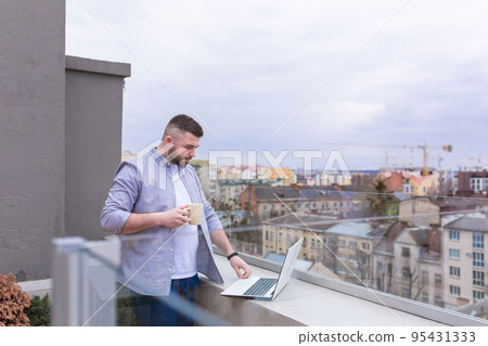 Young man working on the balcony, terrace with laptop, talking on the phone, city view 95431333