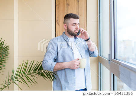 Young man at home talking on the phone, drinking coffee by the window, looking at the camera, smiling, quarantined 95431396