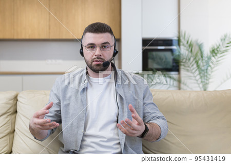 A young man in headphones from home sits on the couch conducts video training business trainings, teaches, explains, looks at the camera A young man in headphones from home sits on the couch conducts video training business trainings, teaches, explains, looks at the camera 95431419