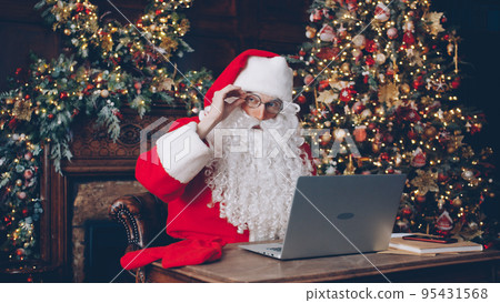 Portrait of thoughtful Santa Claus typing with modern laptop in decorated house using modern device to prepare letters to people. Holidays and surprises concept. 95431568