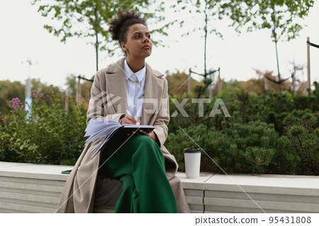 african american woman student making notes in a notebook while drinking coffee outside the office african american woman student making notes in a notebook while drinking coffee outside the office 95431808