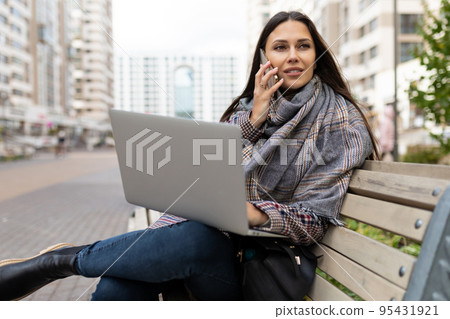 female freelancer speaks on a mobile phone and works on a laptop Sitting on a bench against the female freelancer speaks on a mobile phone and works on a laptop Sitting on a bench against the 95431921