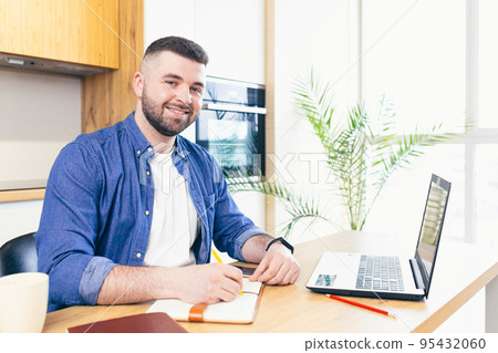young man at home in the kitchen, working on laptop online, talking on the phone, working with documents, making notes, talking video communication, studying, teaching 95432060