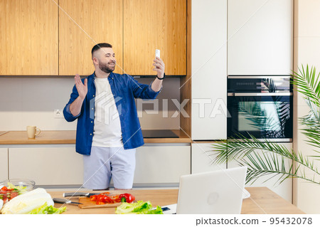A young man at home in the kitchen talking on the phone, on video, learning to cook A young man at home in the kitchen talking on the phone, on video, learning to cook 95432078