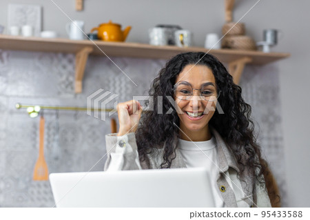 Close up photo of happy and smiling woman looking at laptop screen at home rejoicing and celebrating victory triumph holding hand up, Hispanic woman with curly hair and glasses using netbook. Close up photo of happy and smiling woman looking at laptop screen at home rejoicing and celebrating victory triumph holding hand up, Hispanic woman with curly hair and glasses using netbook. 95433588