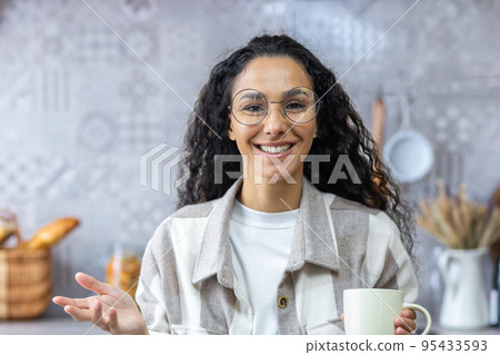 Video call, online communication with friends, happy and smiling hispanic woman looking at web camera and talking, woman at home in kitchen with curly hair and glasses holding a cup of hot drink. Video call, online communication with friends, happy and smiling hispanic woman looking at web camera and talking, woman at home in kitchen with curly hair and glasses holding a cup of hot drink. 95433593