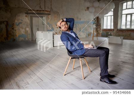 Portrait of businessman sitting on chair in empty room and working on laptop. Making professional analytics and countings on company success Portrait of businessman sitting on chair in empty room and working on laptop. Making professional analytics and countings on company success 95434666