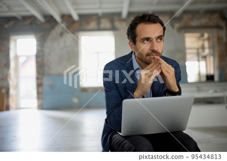 Portrait of man, businessman, worker sitting with laptop with thoughtful expression in big empty room. Professional brainstorming 95434813