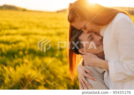 Portrait of loving young couple standing gentle hugging with closed eyes on green meadow in summer evening during golden sunset with warm sunlight. Happy man and woman enjoying time together outdoors. 95435176